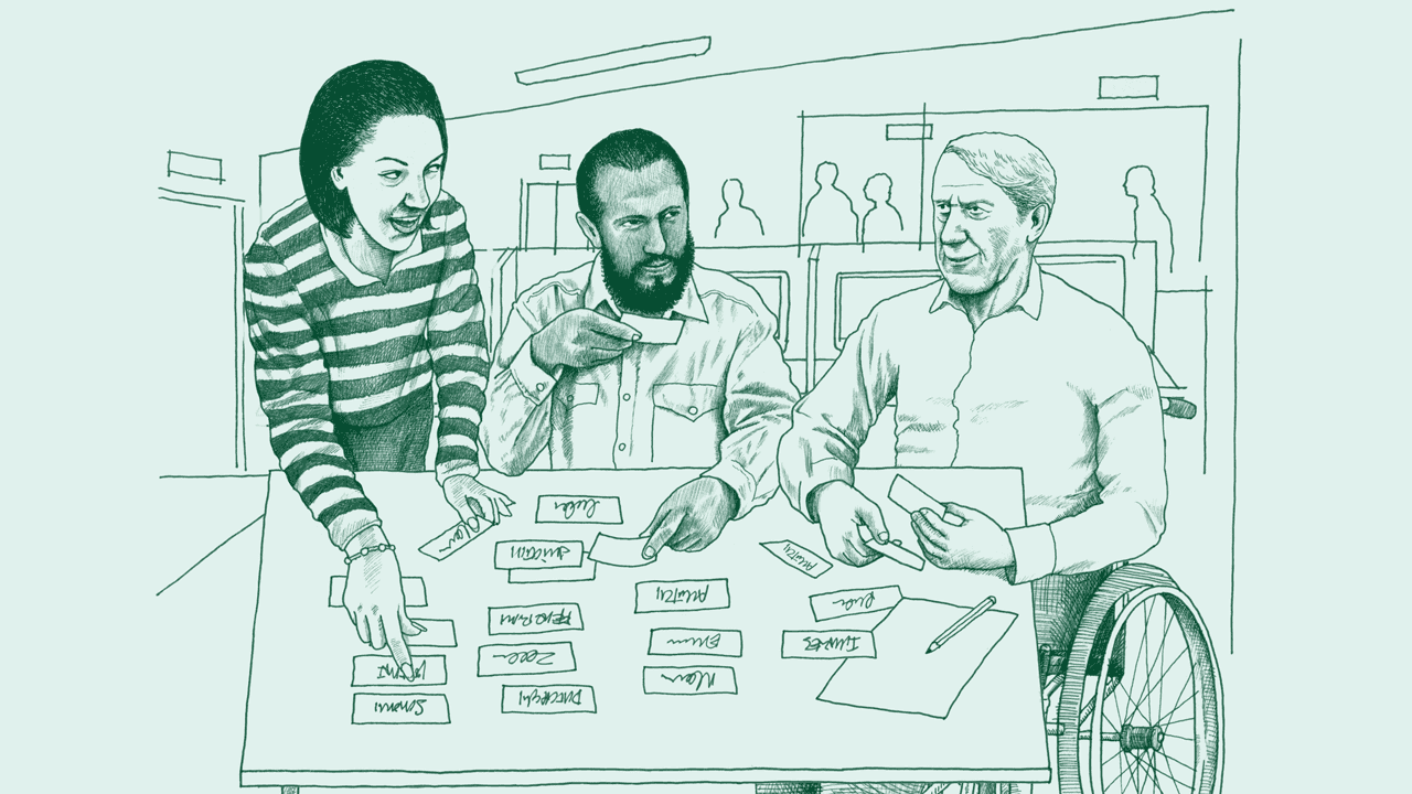 A middle-aged white woman, a middle-aged Middle Eastern man, and an older white man in a wheelchair engage in a card sorting activity at a desk.