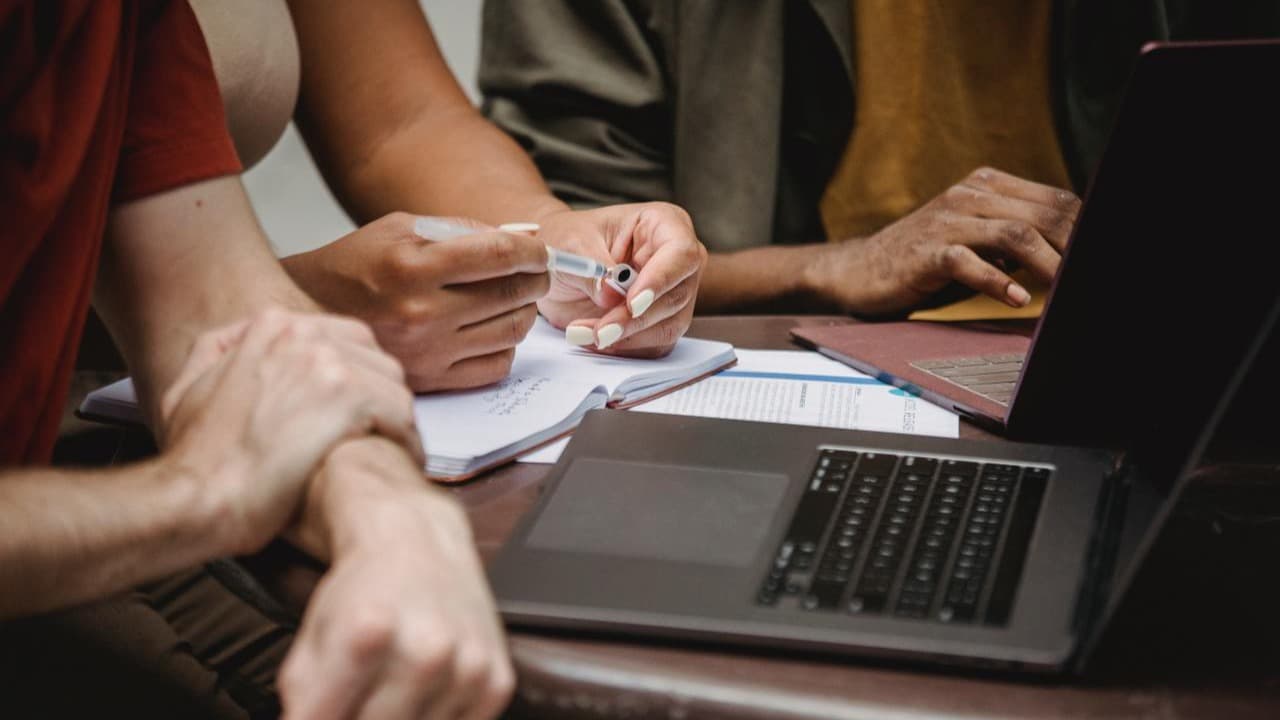 Three people sit at a table with paper, pens, and laptops.