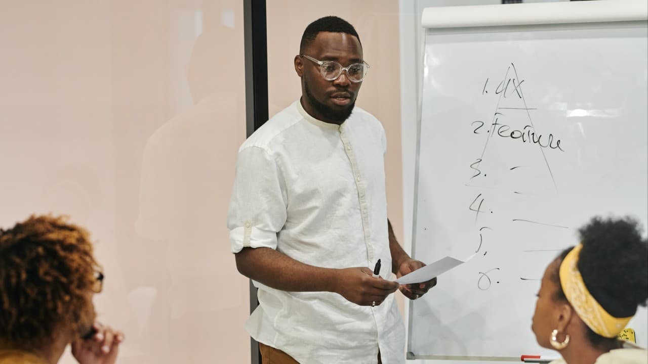 A Black man stands at a whiteboard while in a meeting with his coworkers, two Black women.