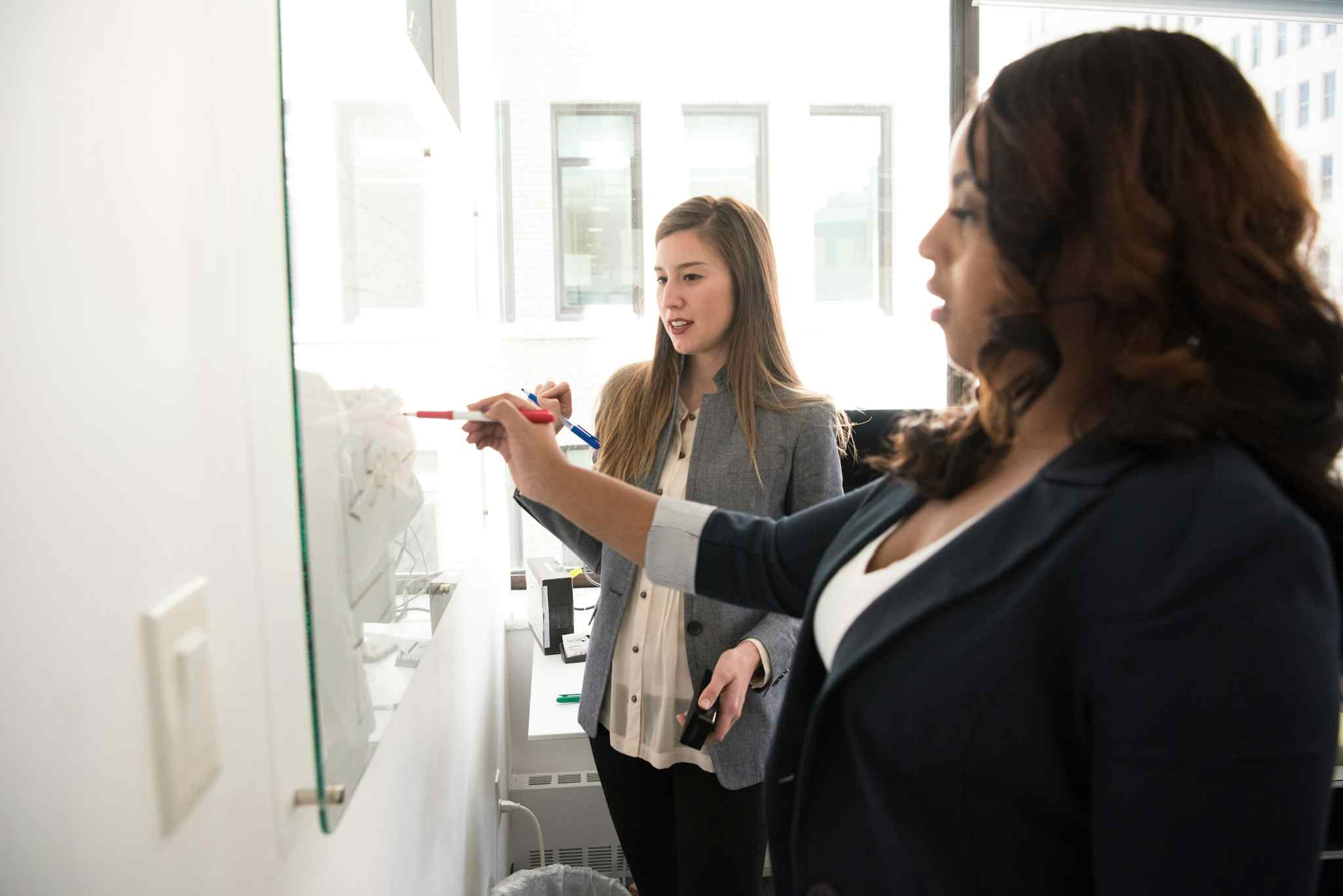 A young Black woman and a young white woman collaborate at a whiteboard.