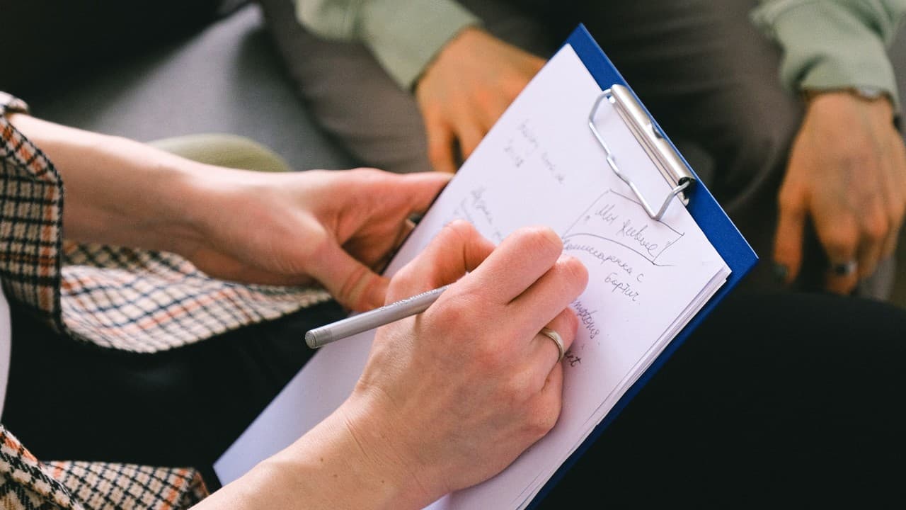 A white woman takes notes on a clipboard while chatting with a white man.