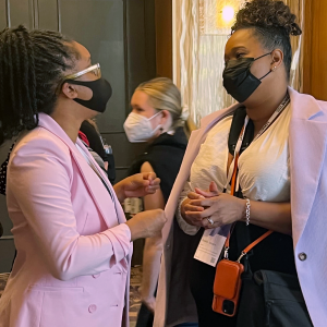 Two Black women wear masks and talk at a conference. The woman on the left works at Nava.