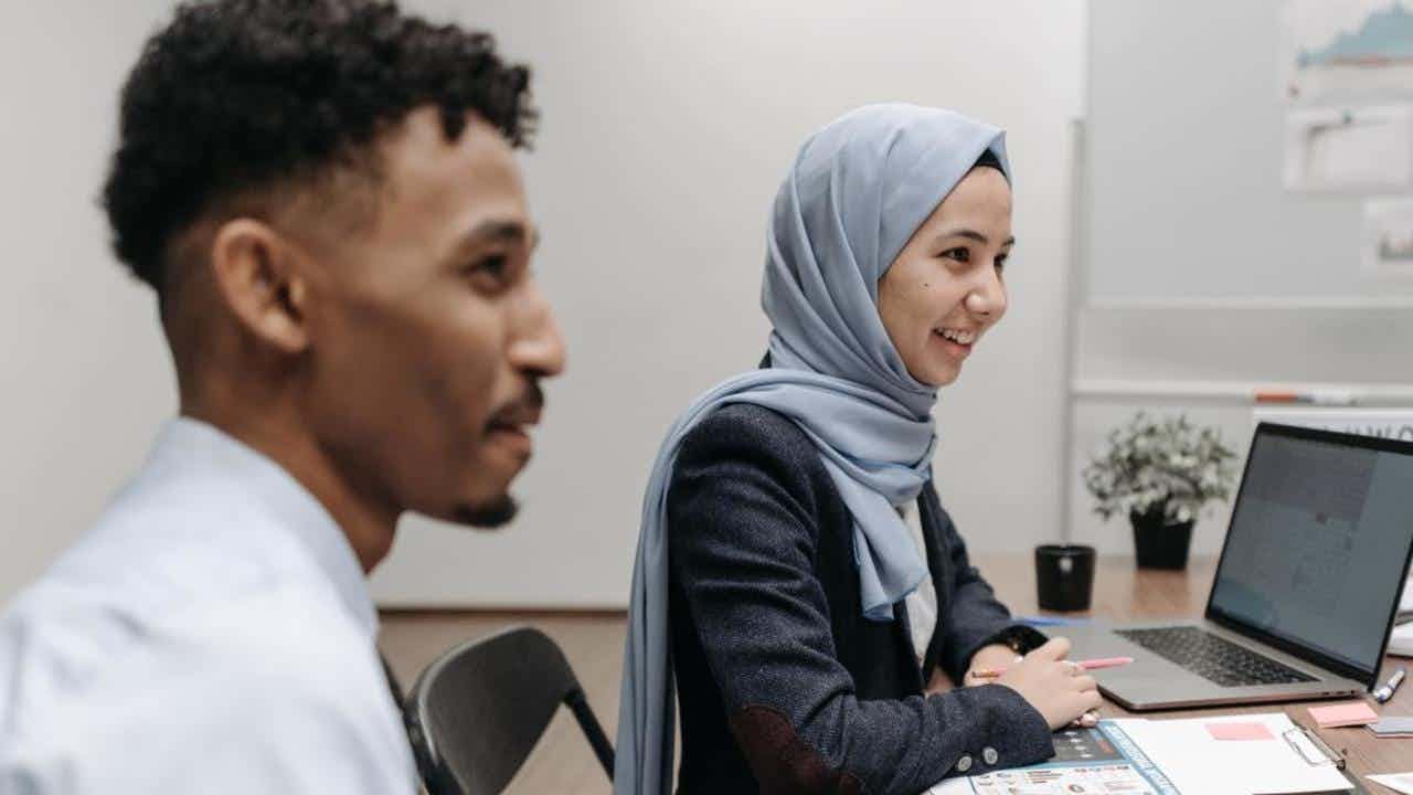 A young Muslim woman wearing a hijab and a young Black man work together on a laptop in an office.