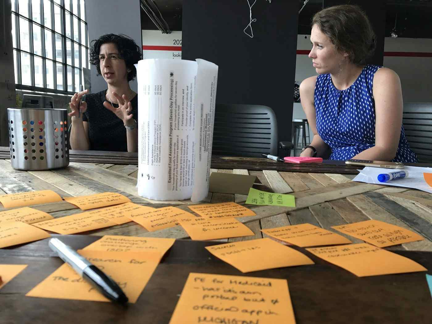 Two white women sit and discuss at a table covered in orange sticky notes.