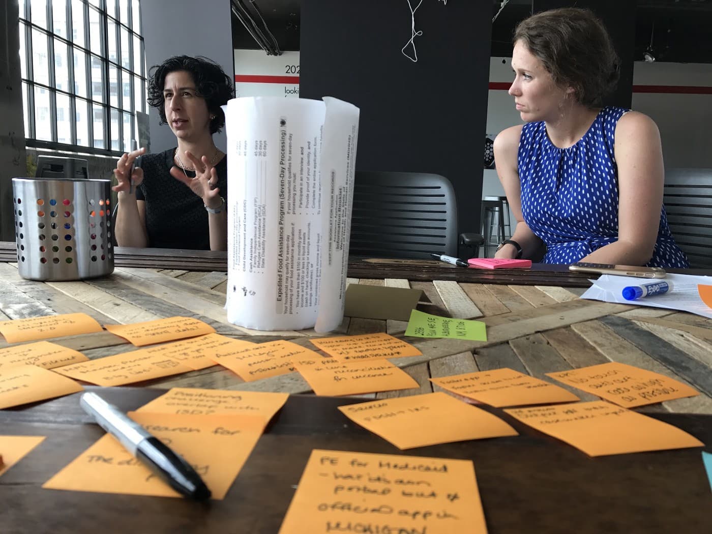 Two white women sit and discuss at a table covered in orange sticky notes.