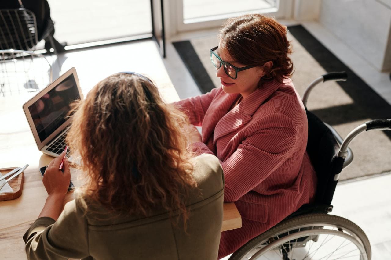 A woman, who wears glasses and uses a wheelchair, is sitting at a table with another woman. The two are looking at a laptop on the table and talking.