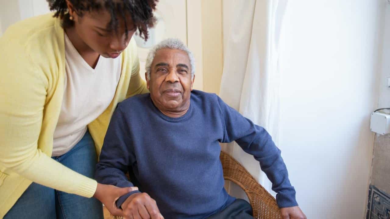 A young Black woman helps an older Black man out of a chair.