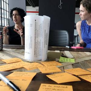 Two white women and Nava employees sit and talk at a conference table covered in sticky notes.