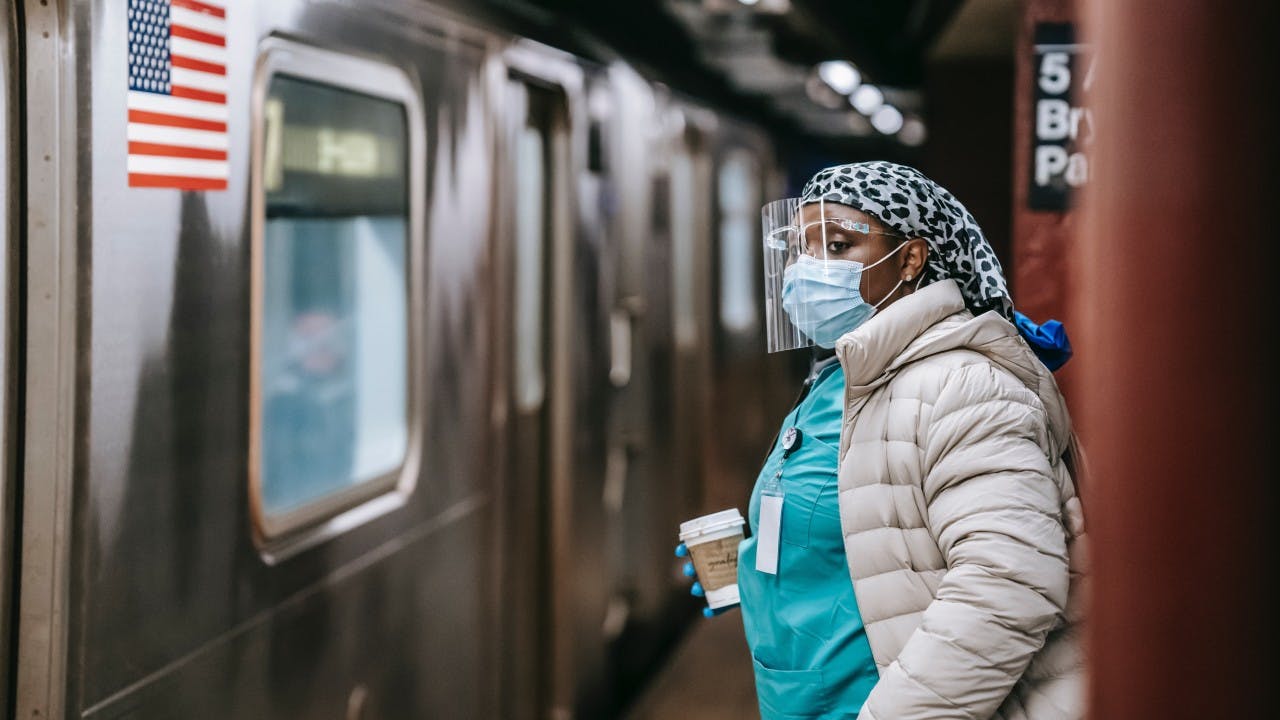 A Black woman wearing scrubs and a mask holds a coffee cup while waiting for her subway train.