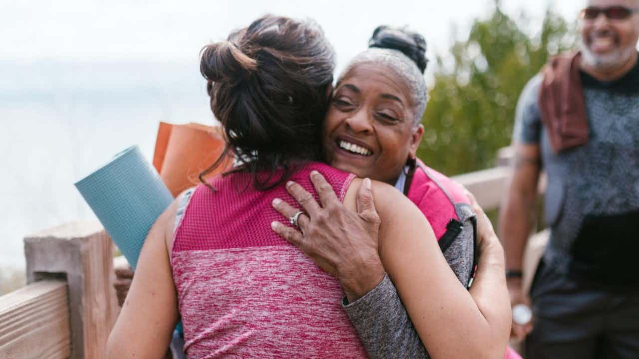 An older black woman hugs a younger white woman. They hold yoga mats. 