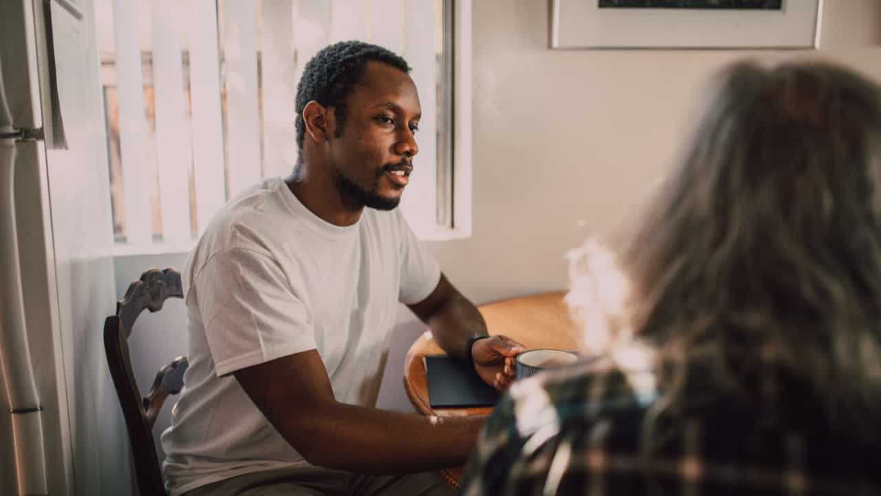 A white man and a Black man talk at a table while drinking coffee.
