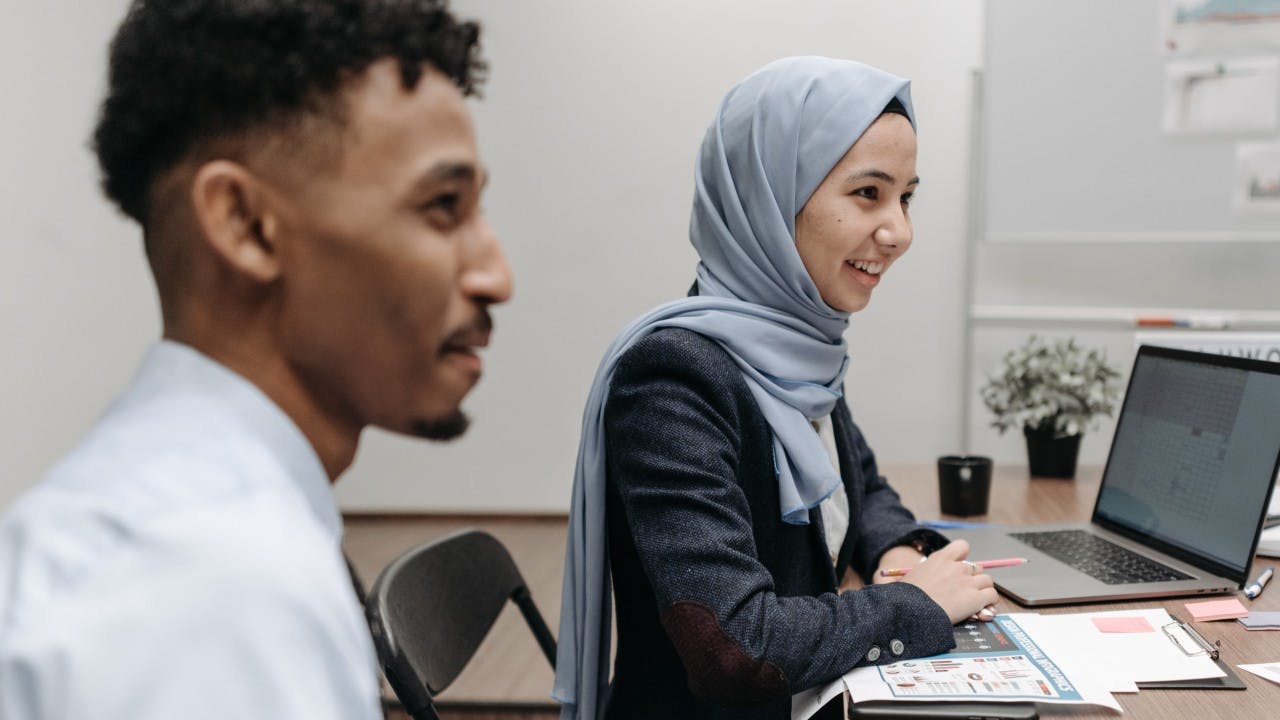 Woman with head scarf smiling in front of a laptop, sitting next to man of color.