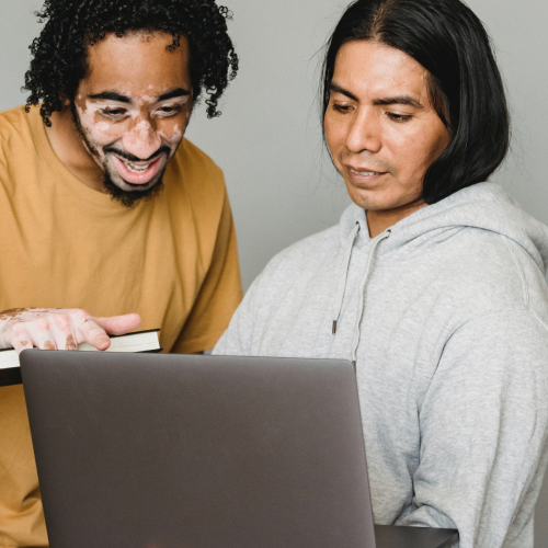 A Black man with vitiligo and an Indigenous American man look at a laptop together.