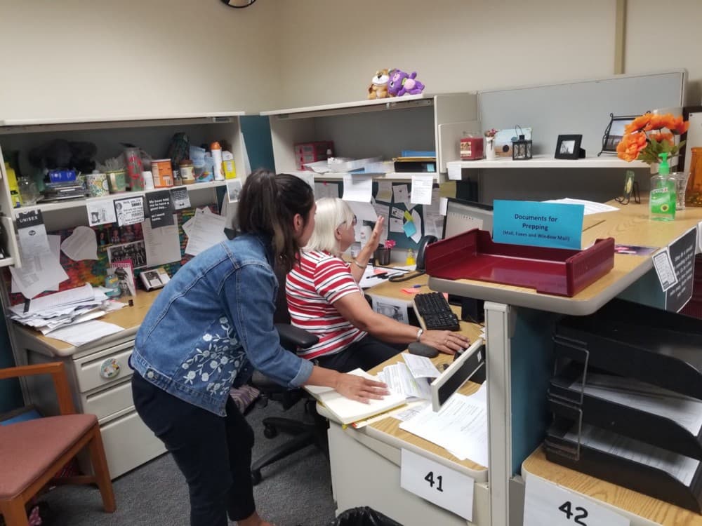 In an office cubicle, a white woman watches and takes notes as another white woman uses her computer.