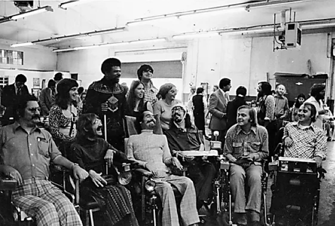 A black and white photo from the 1970s shows a group of people, including some people in wheelchairs, smiling in an office.