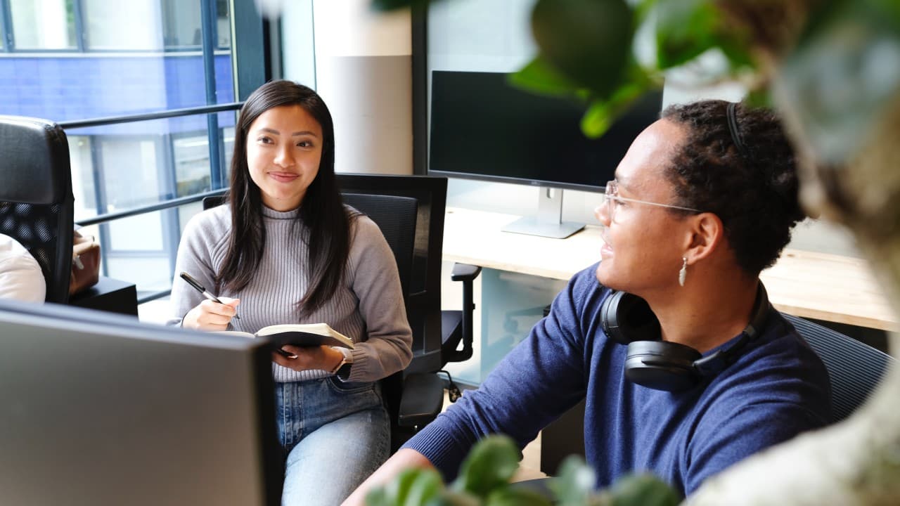 Two coworkers, an Asian woman and a Black woman, take notes while sitting at a computer in an office.
