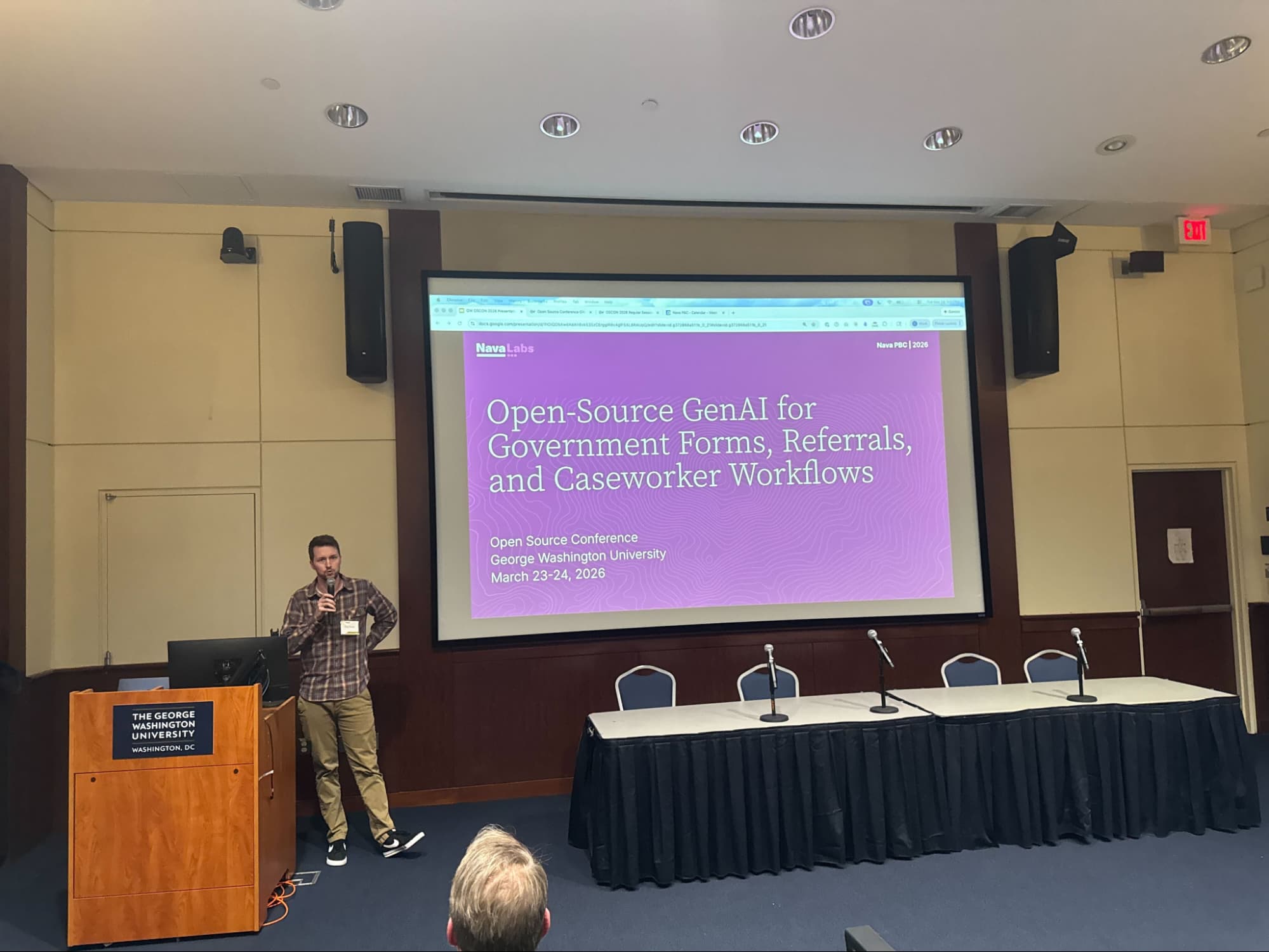 A man stands beside a podium presenting to a group. He stands in front of a screen.