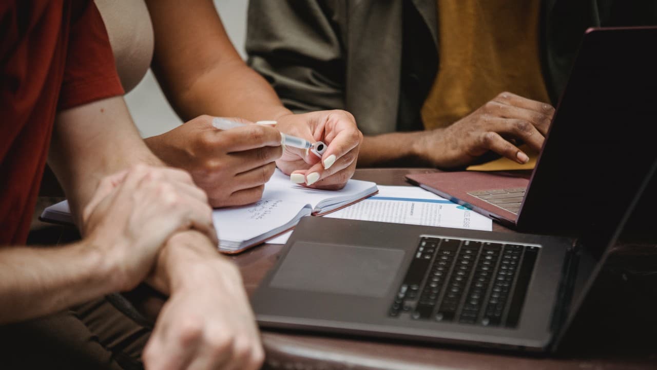 Three people sit around an office table with open notebooks and laptops.