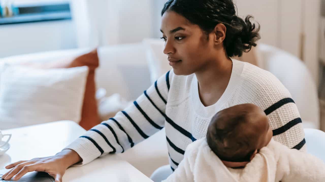 A Black woman holds a baby while using her laptop.